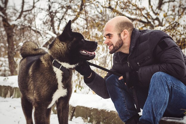 Ein Mann in Jacke und Strickmütze geht mit einem amerikanischen Akita-Hund durch einen verschneiten Wald