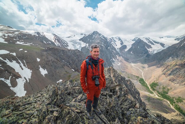 Ein Mann in Rot mit Kamera in der Nähe von Abgrundkante mit scharfen Felsen mit Blick auf große schneebedeckte Berge und Gletscher unter bewölktem Himmel Tourist auf Steinhügel mit Blick auf die Bergkette bei wechselhaftem Wetter