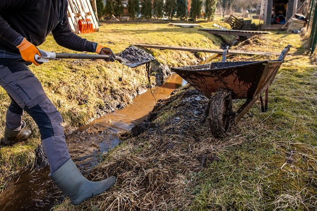 Ein Mann in Schutzausrüstung arbeitet unermüdlich, um einen schlammigen Graben mit einer Schaufel und einer Schlitten in einem Hinterhof im frühen Frühling zu gruben