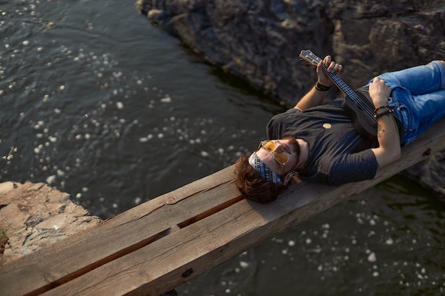 Foto ein mann in schwarzem t-shirt und jeansshorts liegt auf einer holzbrücke über einem bergfluss und spielt den...