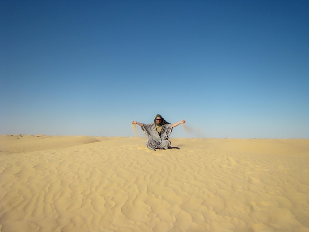 Ein Mann in traditioneller Berbertracht sitzt auf einer Düne in der Wüste Sahara Tunesien Sand spritzt aus seinen Handflächen