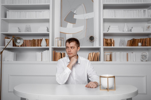 Foto ein mann in weißem hemd sitzt an einem weißen tisch. junger geschäftsmann auf einem schreibtisch. attraktiver geschäftsmann, der am tisch sitzt. vorderansicht eines arbeitenden geschäftsmannes. porträt.