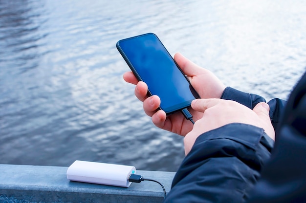 Ein Mann lädt ein Smartphone mit einer Powerbank auf. Das Handy in der Hand wird mit einem tragbaren Ladegerät vor der Kulisse des Sees aufgeladen.