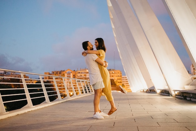 Ein Mann mit Bart umarmt auf einer weißen Brücke in Valencia die Taille seines Mädchens in einem gelben Kleid. Ein paar Touristen in voller Länge bei einem Date am warmen Abend.
