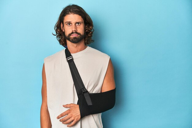 Foto ein mann mit beard, langen haaren und einem arm in einer schleife, der in einem blauen studio schmerzhaft aussieht