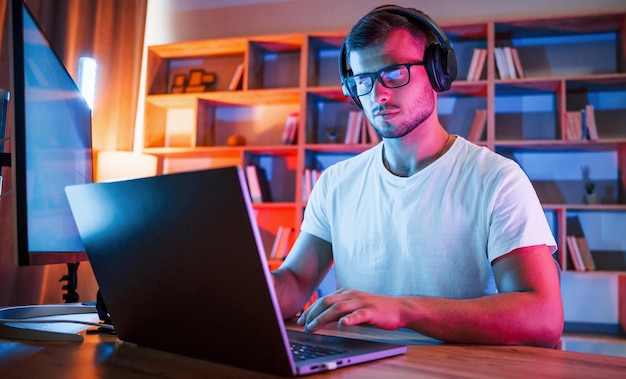 Ein Mann mit Brille und weißem Hemd sitzt neben dem Laptop in einem dunklen Raum mit Neonbeleuchtung