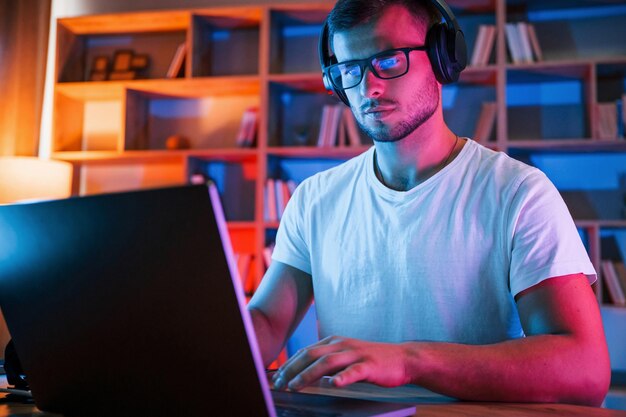 Ein Mann mit Brille und weißem Hemd sitzt neben dem Laptop in einem dunklen Raum mit Neonbeleuchtung