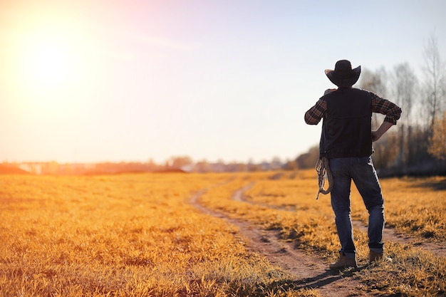Ein Mann mit Cowboyhut und einem Loso auf dem Feld. Amerikanischer Bauer in einem Feld, das einen Jeanshut und mit einem Loso trägt. Ein Mann geht mit Hut über das Feld
