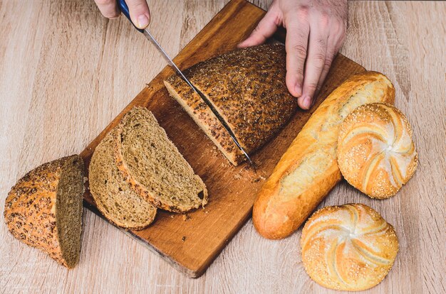 Ein Mann mit einem Messer schneidet Brot auf einem Holzbrett. Französische Baguettes. Verschiedene Rassen auf hölzernem Hintergrund.