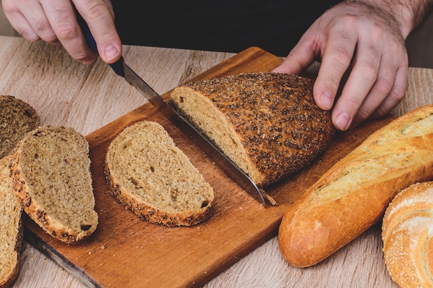 Ein Mann mit einem Messer schneidet Brot auf einem Holzbrett. Französische Baguettes. Verschiedene Rassen auf hölzernem Hintergrund.