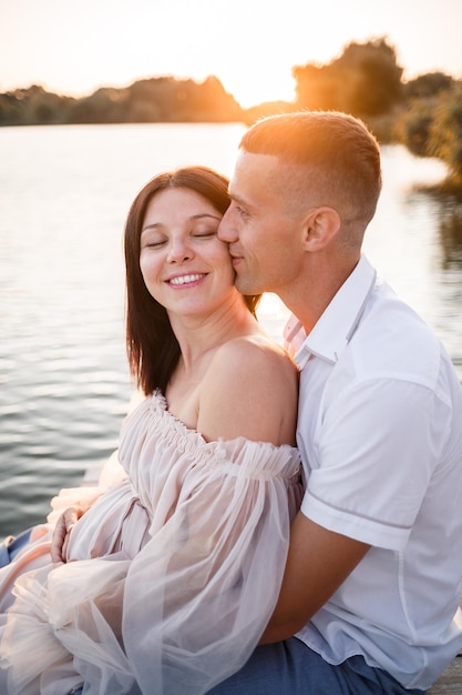 Ein Mann mit einer schwangeren Frau ruht am Ufer des Flusses und sitzt bei sonnigem Sommerwetter bei Sonnenuntergang auf einem Pier Verliebtes Paar Mann und Frau umarmt Händchen haltend