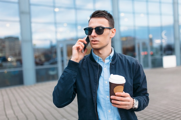 Ein Mann mit einer Tasse Kaffee aus Papier geht durch die Stadt, ein gutaussehender Mann in eleganten Kleidern und Sonnenbrillen, der telefoniert