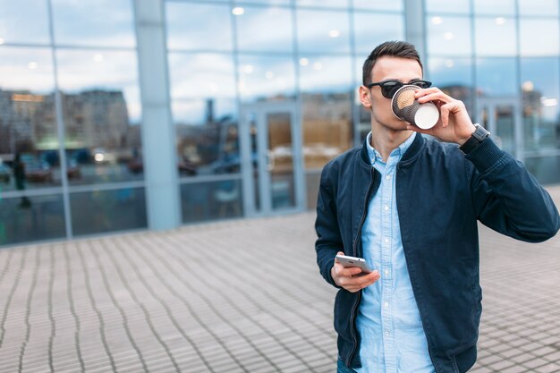 Ein Mann mit einer Tasse Kaffee aus Papier geht durch die Stadt, ein gutaussehender Mann in eleganten Kleidern und Sonnenbrillen, der telefoniert