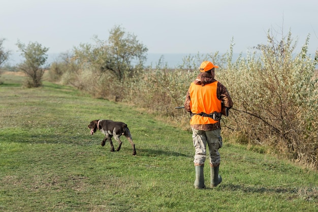 Ein Mann mit einer Waffe in der Hand und einer orangefarbenen Weste auf einer Fasanenjagd in einem Waldgebiet bei bewölktem Wetter Jäger mit Hunden auf der Suche nach Wild