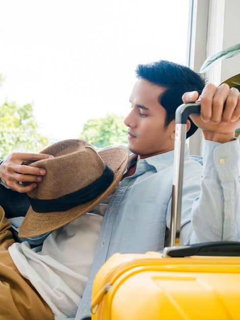 Ein Mann mit gelbem Gepäck Hübscher asiatischer Mann im Jeanshemd, der allein sitzt und wartet und denkt, während er den Koffer in der Nähe des Glasfensters im vertikalen Stil hält Sommerurlaubsreise-Urlaubskonzept