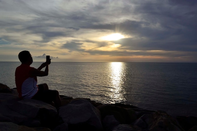 Ein Mann mit Gesichtsmaske fotografiert mit einem Smartphone den wunderschönen Sonnenuntergang am Strand Tanjung Aru Kota Kinabalu in Borneo Sabah Malaysia