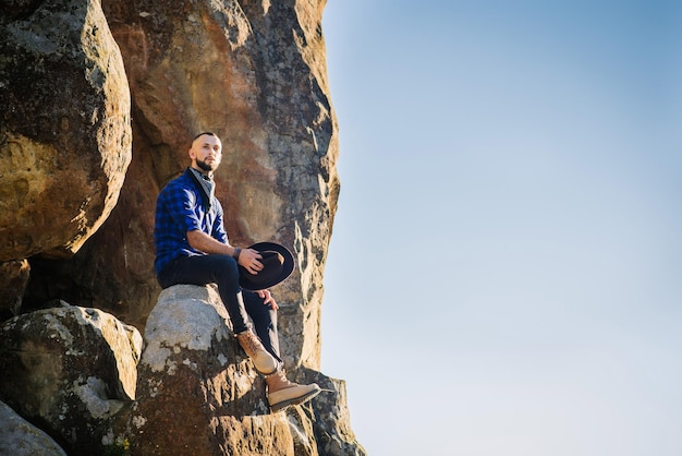 Ein Mann mit Hut, der hoch auf den Felsen am blauen Himmelshintergrund sitzt