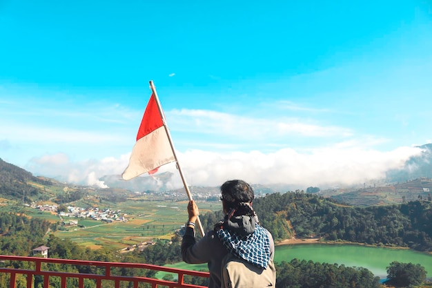 Ein Mann mit indonesischer Flagge, der auf einer Klippe steht und den Blick auf die Natur von See und Berg in Dieng Indonesien genießt