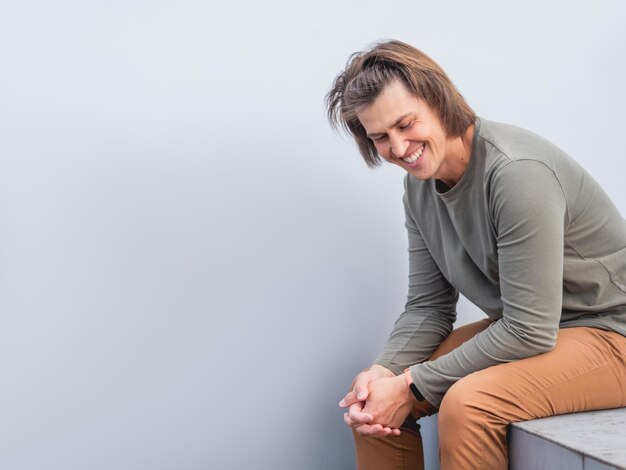 Foto ein mann mit langen haaren und einem glücklichen aussehen sitzt in der nähe einer grauen wand und lächelt. ein mann in leichter kleidung.