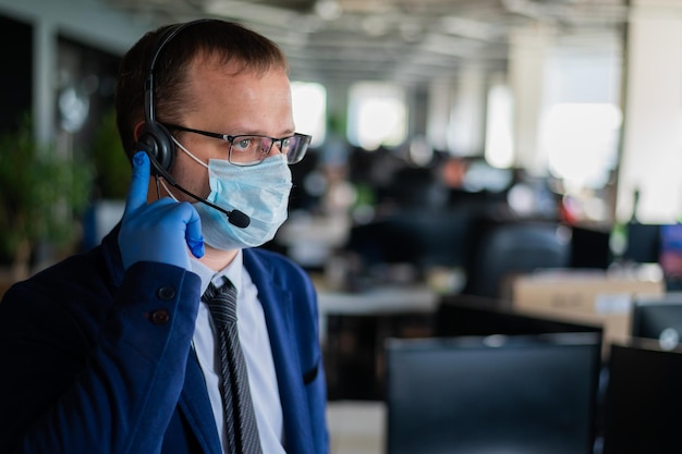 Foto ein mann mit latexhandschuhen und einer medizinischen maske spricht über das headset mit einem kunden. ein männlicher büroleiter beantwortet anrufe. arbeit während des coronavirus. krawatte und sakko eines angestellten