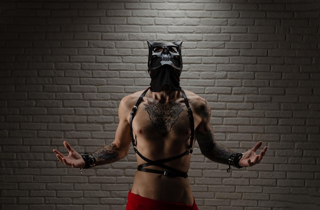Ein Mann mit nacktem Oberkörper in einer BDSM-Dämonenmaske mit Tätowierungen und Ledergürteln und Handschellen