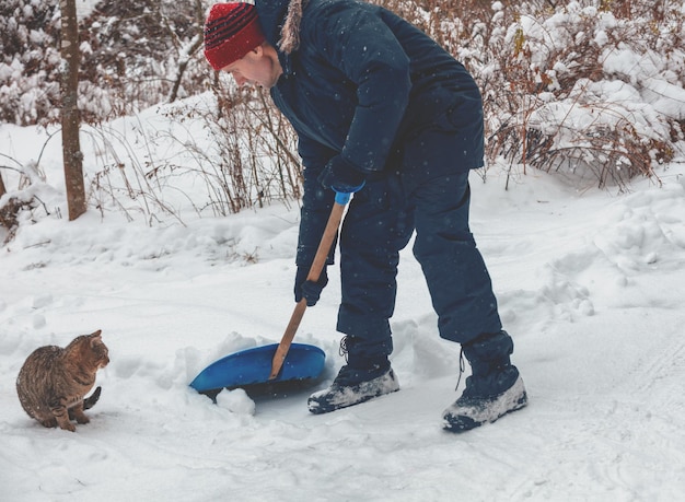 Ein Mann mit Schaufel reinigt die Straße bei Schneefall vom Schnee. Katze sitzt in der Nähe