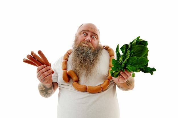Ein Mann mit schwerem Gewicht und Würstchen um den Hals hat Schwierigkeiten bei der Auswahl von Lebensmitteln: Würstchen oder Spinat. Porträt lokalisiert auf weißem Hintergrund