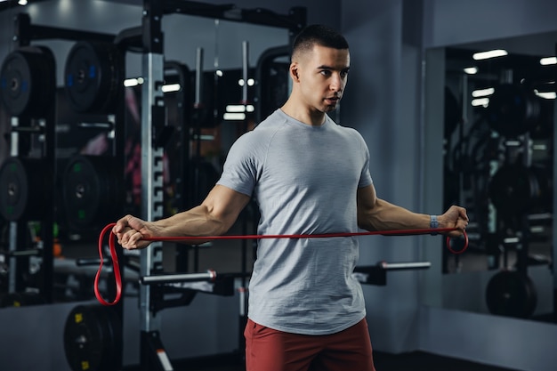 Ein Mann mit sichtbaren Adern an den Armen, der mit einem Gummiband in einem Fitnessstudio mit großen Spiegeln und schwarzen Matten trainiert