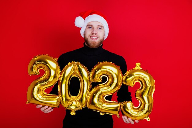 Ein Mann mit Weihnachtsmannmütze hält die glänzenden Zahlen des neuen Jahres 2023 auf rotem Grund