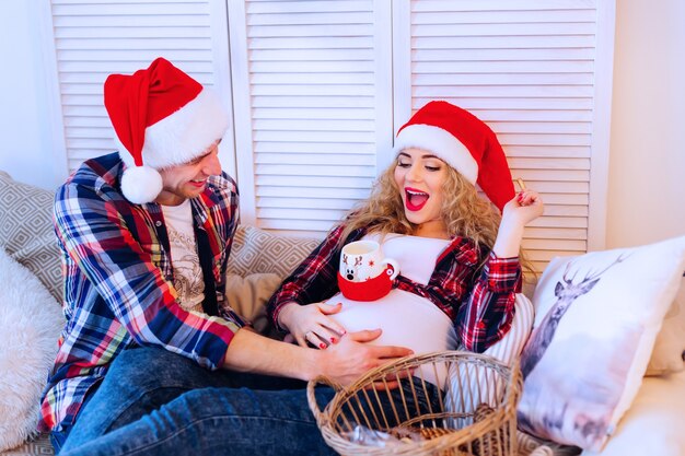 Ein Mann mit Weihnachtsmannmütze und seine schwangere Frau sitzen auf der Couch