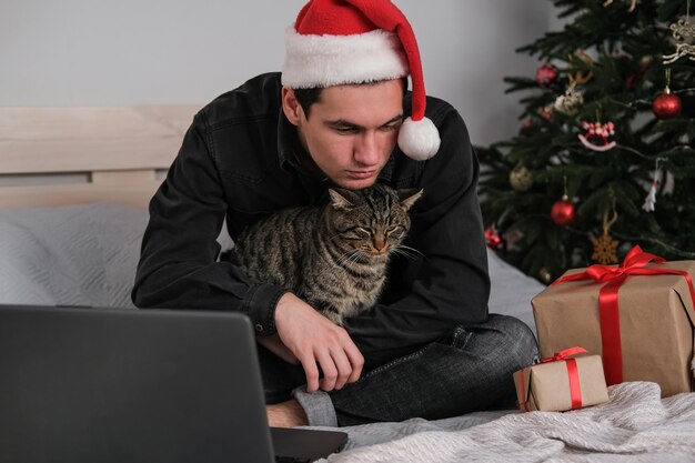 Ein Mann mit Weihnachtsmütze sitzt auf einem Bett und umarmt eine Katze und ist traurig, dass er alleine Weihnachten feiert