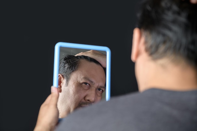 Ein Mann mittleren Alters glänzt im Spiegel und sorgt sich um Haarausfall oder Haargrau isoliert auf schwarzem Hintergrund Gesundheitskonzept