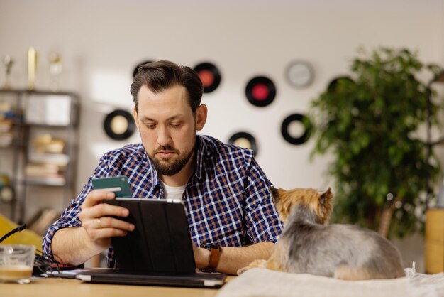 Ein Mann mittleren Alters sitzt an seinem Schreibtisch und hält ein Tablet und eine Zahlungskarte in der Hand, mit der er sich in sein Unternehmen einloggt