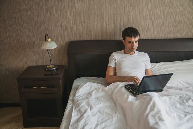 Ein Mann mittleren Alters sitzt in seinem Schlafzimmer im Bett und arbeitet an einem Laptop. Das Konzept der Remote-Arbeit