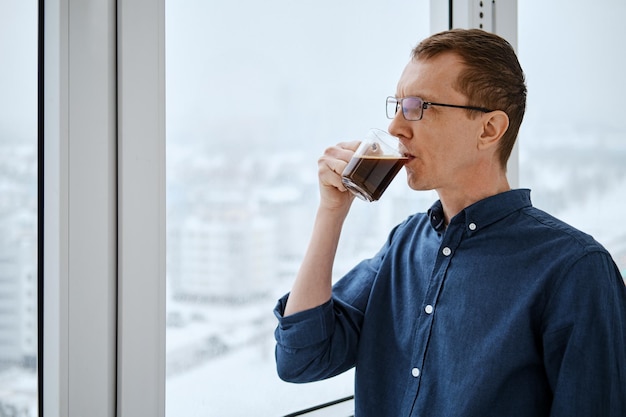 Ein Mann mittleren Alters steht am frühen Wintermorgen auf dem Balkon und trinkt Kaffee
