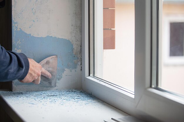 ein mann repariert zu hause, in seinen händen einen spachtel, mit dem er alte farbe von den wänden reinigt