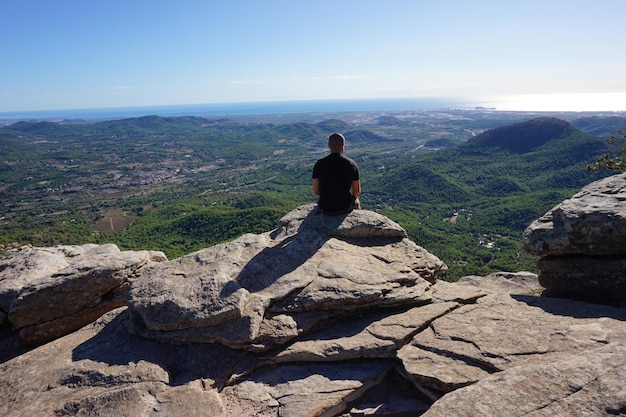 Ein Mann sitzt auf einem Felsen mit Blick auf ein Tal und das Meer.