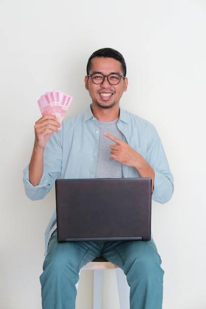 Ein Mann sitzt auf einer Bank mit einem Laptop auf dem Schoß und hält mit einem glücklichen Gesichtsausdruck Geld