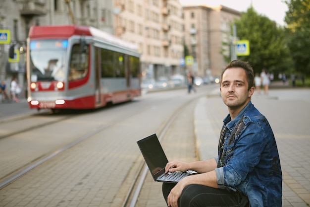 Ein Mann sitzt mit einem Laptop auf dem Schoß in der Nähe der Straßenbahnlinie.