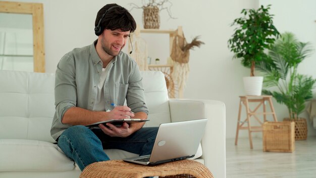 Ein Mann sitzt mit einem Laptop und einem Headset auf einem Sofa im Wohnzimmer des Hauses und kommuniziert über eine Videoverbindung.