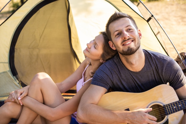 Ein Mann spielt für eine Frau auf einer Gitarre in der Nähe eines Zeltes bei Sonnenuntergang. Flitterwochen in der Wanderung