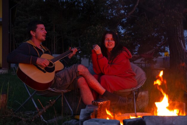 Ein Mann spielt Gitarre, eine Frau hört zu und singt mit. Ein verliebtes Paar sitzt am Lagerfeuer im Innenhof des Hauses auf Campingstühlen, ein romantischer Abend.