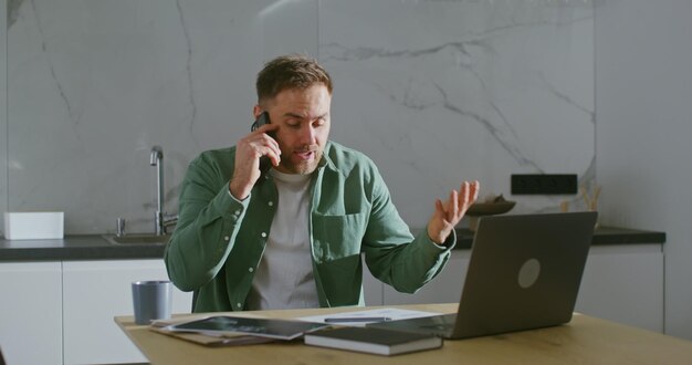 Ein Mann spricht am Handy und sitzt am Küchentisch vor dem Laptop