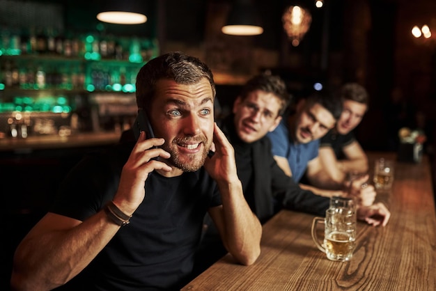 Ein Mann spricht am Telefon, drei Sportfans in einer Bar schauen Fußball mit Bier in der Hand.