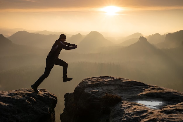 Ein Mann springt bei Sonnenuntergang über den Gipfel eines Gipfels und macht einen Glaubenssprung von einem Felsvorsprung