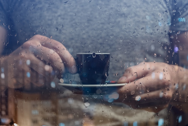Ein Mann steht am Fenster und trinkt Kaffee. Regentropfen auf Glas.