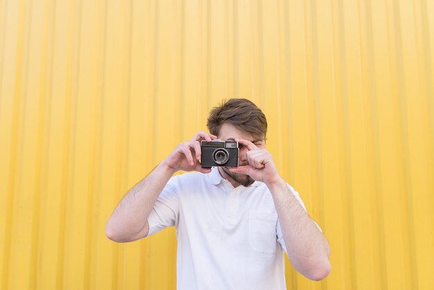Ein Mann steht an einer gelben Wand und macht ein Foto auf einer Retro-Kamera.