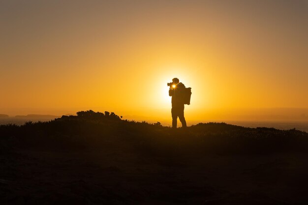 Ein Mann steht auf einer Klippe und fotografiert das Meer bei Sonnenuntergang