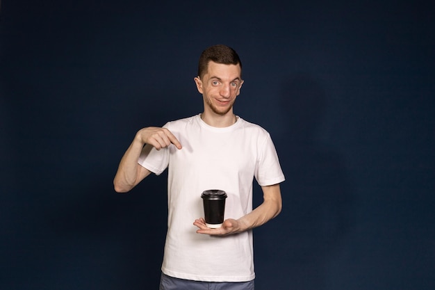 Ein Mann steht vor blauem Hintergrund und hält einen Kaffee in der Hand