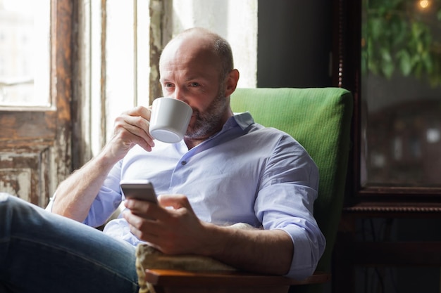 Ein Mann trinkt Kaffee und überprüft die Nachrichten im Telefon, während er auf einem Stuhl am Fenster sitzt